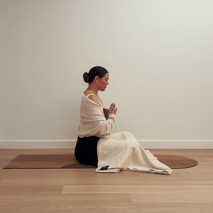 Woman meditating in a prayer pose on a brown yoga mat with a white blanket.