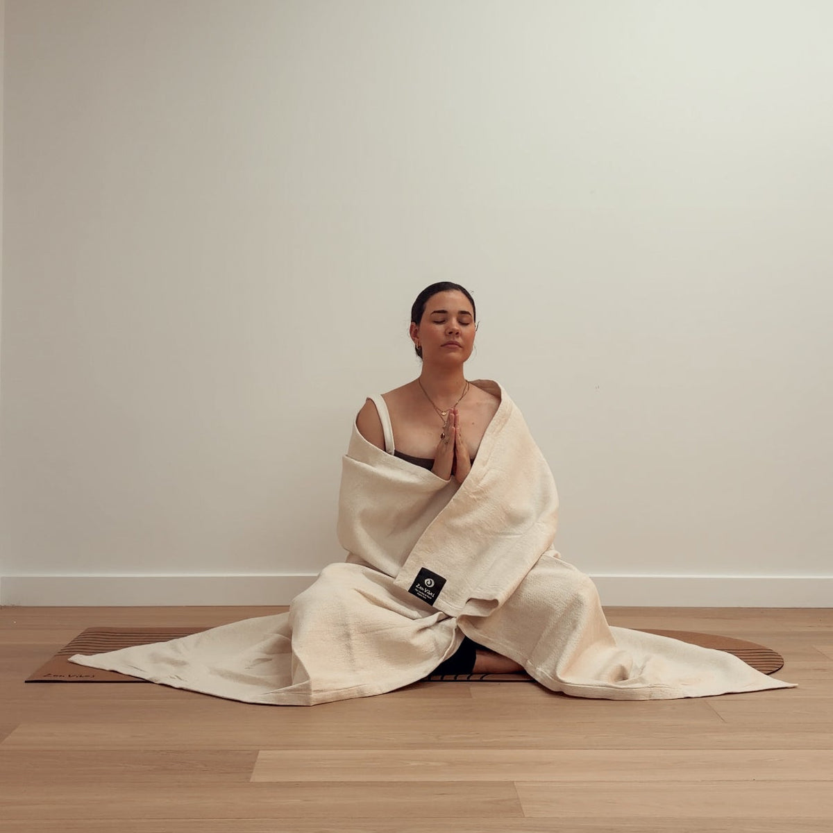 Woman meditating in a prayer pose wrapped in a cream-colored yoga blanket on a mat.
