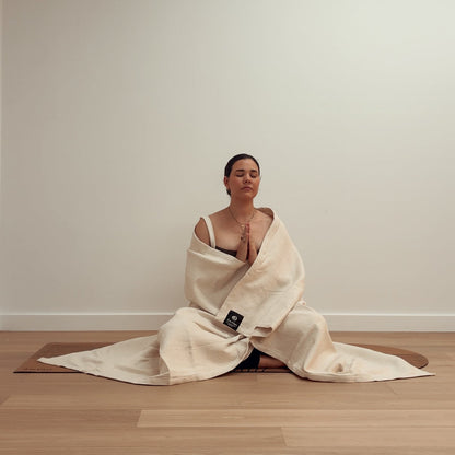Woman meditating in a prayer pose wrapped in a cream-colored yoga blanket on a mat.